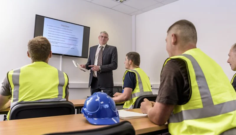 A trainer presents course objectives on a screen to workers in hi-vis vests in a classroom
